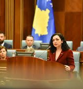 A female politician or government official delivers a speech from a podium in a formal parliamentary chamber or meeting room